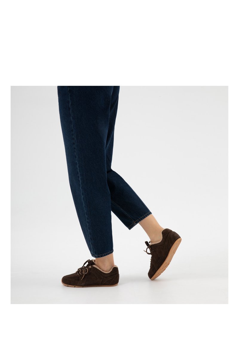 Persona che indossa jeans blu scuro e sneaker marroni foderate in pelliccia con suole di gomma, sollevando un piede su uno sfondo bianco semplice.