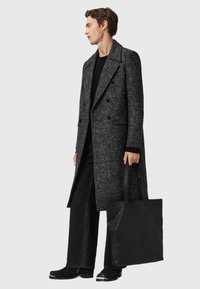 Grå tweed frakke med dobbeltradet design, båret over en sort sweater og sorte brede bukser. Sort tote bag med teksturerede accenter.