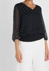 Svart blus med V-ringning och trekvartslång ärm. Har ett geometriskt silvermönster och elastisk midja. Bärs tillsammans med vita byxor.