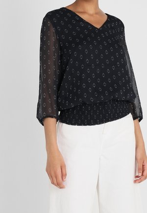 Blusa nera con scollatura a V e maniche a tre quarti. Presenta un motivo geometrico argento e una fascia elastica in vita. Indossata con pantaloni bianchi.
