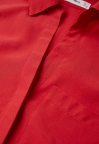 Primo piano di una camicia rossa a maniche lunghe che mostra il colletto, la patta e una tasca sul petto con dettagli di cucitura visibili.