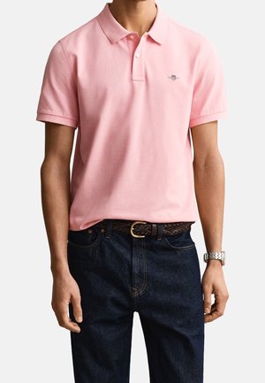 Homme portant un polo rose clair à manches courtes, un jean bleu foncé avec une ceinture marron tressée et une montre-bracelet argentée au poignet gauche.