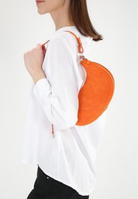 Okrągła, pomarańczowa torebka z zamszu o zakrzywionej sylwetce, cienkim pasku i szczególe zamka, noszona na tle białej koszuli. Prosty, nowoczesny design.