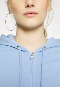 Fleece azul claro com fecho éclair, apresentando um toque suave, com hardware em tom prateado e cordões, usado com grandes brincos de argola prateados.