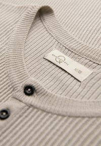 Maglione beige a coste con collo rotondo, caratterizzato da bottoni neri e un'etichetta tessile con informazioni sul marchio e sulla taglia.