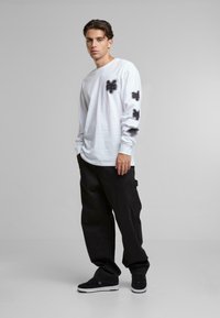 White long-sleeve shirt with black spray-paint-style designs on the chest and sleeve, paired with loose-fitting black pants and black sneakers.