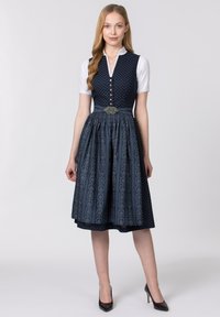 Dunkelblaues Dirndlkleid mit gemustertem Rock, geknöpftem Oberteil und taillierter Taille, verziert mit einer dekorativen silbernen Brosche. Darunter eine weiße Bluse.