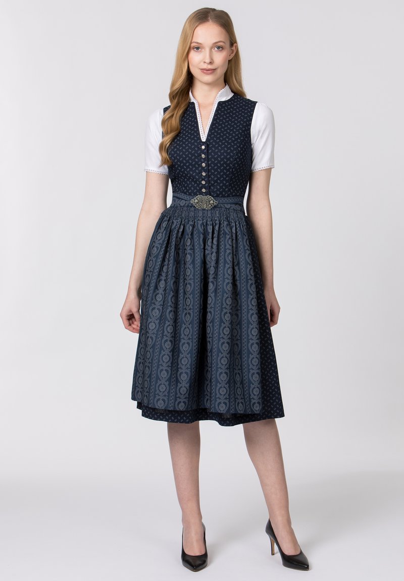 Dunkelblaues Dirndlkleid mit gemustertem Rock, geknöpftem Oberteil und taillierter Taille, verziert mit einer dekorativen silbernen Brosche. Darunter eine weiße Bluse.