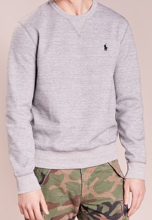 Person trägt ein hellgraues Sweatshirt mit langen Ärmeln, kleinem schwarzem Logo und grünen Camouflage-Cargohosen.