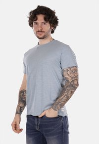T-shirt azzurro chiaro a maniche corte in tessuto morbido, con scollatura rotonda e vestibilità rilassata. Dettagli di tatuaggi distintivi visibili sulle braccia.