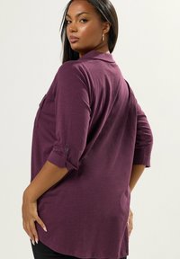 Camicia a maniche lunghe e dal taglio morbido in tessuto viola scuro. Presenta polsini con bottoni e una trama delicata. Le fessure laterali aggiungono movimento all'orlo.