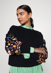 Μαύρος πουλόβερ με φουσκωτά μανίκια κεντημένα με λουλούδια, που διαθέτει πολύχρωμα λουλούδια. Κάτω από αυτό, είναι ορατό ένα ανοιχτό πράσινο πουκάμισο με γιακά.