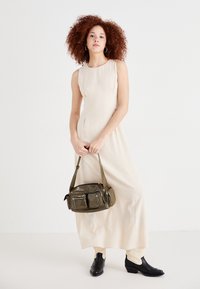 Robe sans manches beige avec des plis latéraux, associée à un sac vert foncé avec des poches zippées et une bandoulière réglable, ainsi qu'à des bottines noires.