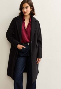 Svart ullkappa med slagkrage, enkel knappstängning och sidofickor. Bärs över en röd blus och kombineras med mörkblå jeans.