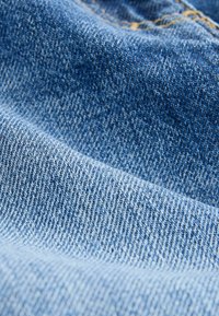 Denimstoff in einem Farbverlauf von Blautönen, mit feiner Webart und leicht strukturierter Oberfläche. Sichtbare Nähte in kontrastierendem Gelb.