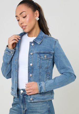 Junge Frau trägt eine hellblaue Jeansjacke und Jeans, richtet den Jackenkragen und schaut leicht nach links vor schlichtem Hintergrund.