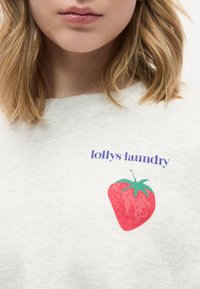 Κοντινό πλάνο του κάτω μέρους του προσώπου και του λαιμού ενός ατόμου που φοράει ανοιχτό γκρι πουκάμισο με μοβ κείμενο «lollys laundry» και κόκκινο γραφικό φράουλας.