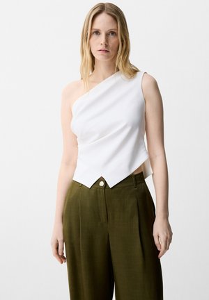 Femme blonde portant un top blanc asymétrique sans manches et un pantalon large taille haute vert olive, debout devant un fond blanc.