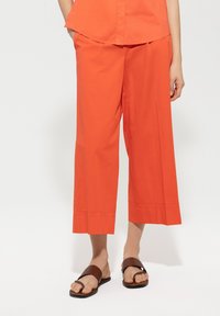 Orange wide-leg trousers made of lightweight fabric, featuring a pleated waistband and a cropped length, paired with brown flat sandals.