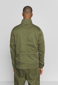 Veste légère vert olive avec col montant, taille élastique et détail de plastron au dos ; présente des coutures contrastées et des manches longues.