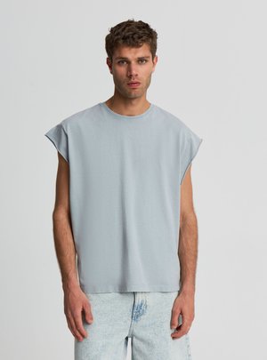 Giovane uomo con capelli corti e ricci che indossa una canotta grigio chiaro e jeans di denim azzurri chiari su uno sfondo grigio uniforme.