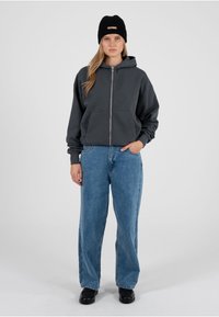 Grauer Kapuzenpullover mit Reißverschluss und Fronttaschen, kombiniert mit weiten blauen Jeans und schwarzen Turnschuhen. Das Modell trägt eine schwarze gerippte Mütze. Einfaches Design.