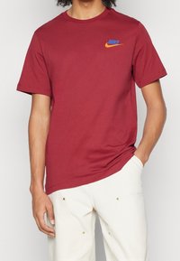 Bordowa bawełniana koszulka z krótkim rękawem i okrągłym dekoltem. Małe niebiesko-pomarańczowe logo Nike po lewej stronie na klatce piersiowej. Prosty design, luźny fason.
