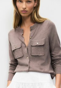 Hnědý kardigan s výstřihem henley, dvěma předními kapsami se klopami a knoflíky ve zlatém odstínu. Tkanina se zdá být měkká s hladkou texturou.