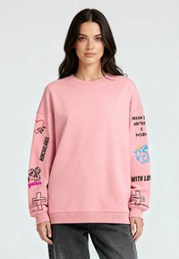 Világos rózsaszín pulóver hosszú ujjal, kerek nyakkal, fekete és kék grafikákkal az ujjakon, és bordázott mandzsettával az alján.