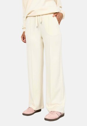 Person trägt cremefarbene Hosen mit weitem Bein und Kordelzug in der Taille sowie rosa Wildledersneaker, Hand in der Tasche, vor einfachem Hintergrund.