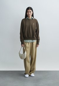 Bruine oversized trui, witte overhemd met groene strepen, beige wijde broek en witte sneakers, gecombineerd met een kleine crèmekleurige schoudertas.