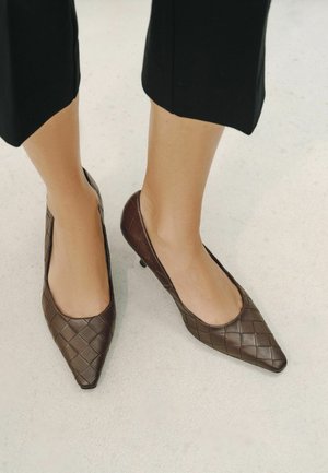 Donna che indossa scarpe intrecciate marrone scuro con punta appuntita e tacco basso con pantaloni neri corti su un pavimento chiaro.