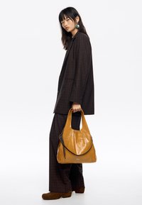 Gran bolso de cuero color mostaza con textura brillante, que presenta una correa de acento negra. El modelo lleva un traje de cuadros oscuros y botas marrones.