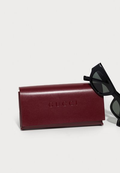 Burgundi nahast Gucci prillikarp mustade niitidega cat-eye prillide kõrval, millel on tumedad läätsed, valgel taustal.