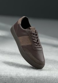 Baskets en cuir marron avec des accents en daim, présentant un design perforé, des lacets et une semelle en caoutchouc texturée. Nom de la marque visible sur le talon.