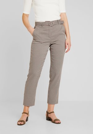 Vrouw draagt een broek met hoge taille en geruite print met riem, een wit geribd topje en bruine sandalen met hak tegen een effen achtergrond.