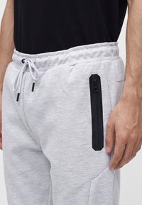 Uomo che indossa pantaloni della tuta grigio chiaro con tasca verticale nera con cerniera e coulisse bianca, in piedi con la mano rilassata al fianco.