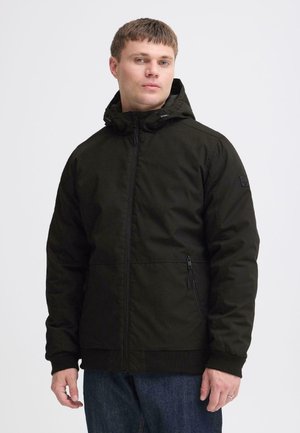 Homme portant une veste noire à capuche avec des poches zippées, debout devant un fond blanc uni.
