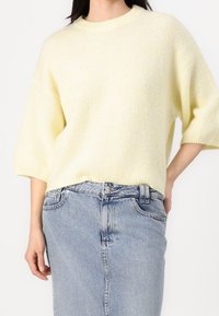 Helles gelbes Strickpullover mit kurzen, weiten Ärmeln und geripptem Halsausschnitt, kombiniert mit einem blauen Jeansrock. Weiche Textur und lockere Passform.