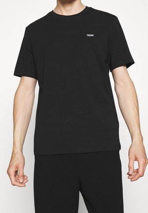 Mężczyzna ubrany w czarny T-shirt z krótkim rękawem i okrągłym dekoltem oraz czarne spodnie, z małym białym logo "HUGO" na lewej piersi.