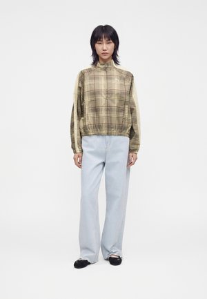 Person iført en grøn ternet lynlåstrøje, lyseblå vidde jeans og sorte sko, der står mod en ensfarvet hvid baggrund.