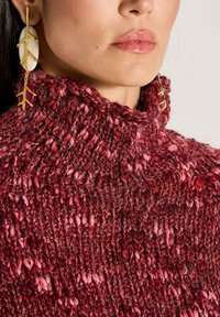 Vrouw in een chunky rode en roze gebreide coltrui met gouden oorknoppen in de vorm van bladeren en witte stenen.