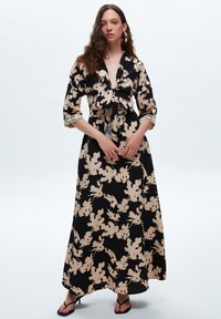 Abito lungo in tessuto nero con motivo floreale beige. Presenta una parte anteriore annodata, maniche 3/4 con dettagli in pizzo e una silhouette fluida.
