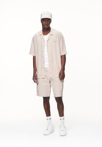 Beige kortærmet skjorte med knapper over en hvid tanktop, parret med beige cargo shorts. Accessories inkluderer en lys farvet kasket og hvide sneakers.
