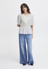 Haut peplum blanc à manches courtes avec boutonnage à l'avant, décolleté en V et taille froncée, associé à un jean clair à jambes larges et des chaussures noires.