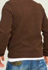 Chaqueta de forro polar marrón con una superficie texturizada, con mangas largas y un dobladillo recto. Usada sobre una camisa blanca y unos vaqueros.