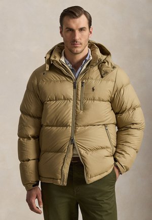 Uomo che indossa una giacca imbottita color sabbia con cappuccio sopra una camicia a righe, in piedi davanti a uno sfondo beige uniforme, mani in tasca.