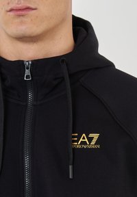 Felpa nera con zip e colletto alto, dotata di un logo dorato EA7 Emporio Armani sul lato sinistro del petto. Tessuto testurizzato con cordino.