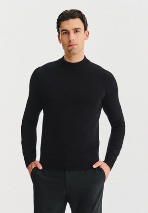 Czarny dzianinowy sweter z golfem z długimi rękawami, dopasowanym fasonem i ściągaczami na mankietach. Noszony na co dzień z rękami w kieszeniach, w połączeniu z ciemnymi spodniami.