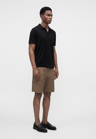 Homme debout portant un polo noir à manches courtes, un short marron aux genoux et des mocassins noirs sur un fond clair uni.